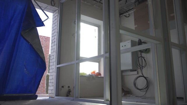 Interior construction site shows metal wall frames and electrical wiring. Blue tarp covers doorway in unfinished room with bright window