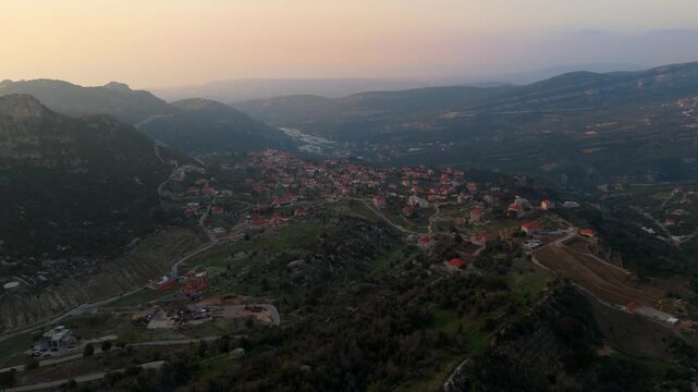 Drone footage capturing Douma village spread across the mountain landscape at sunset, with winding roads, red-roofed houses, and layered hills in Lebanon.