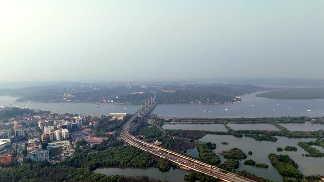 Cinematic aerial view of Atal Setu spanning across the Mandovi River, with calm waters, scattered boats, and distant coastal settlements fading into a hazy horizon.