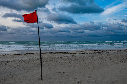Red warning flag standing tall on a beach with cloudy skies and waves