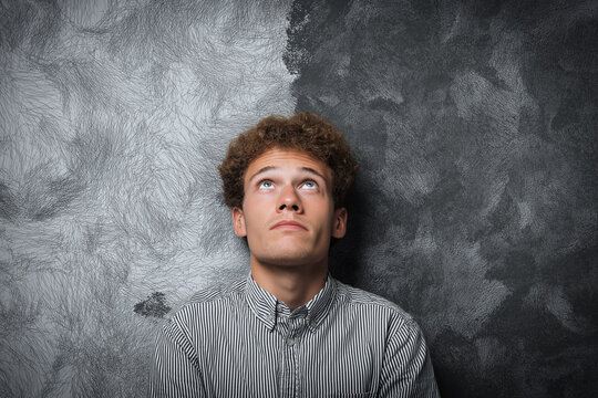 A young man with curly brown hair, wearing a striped button-up shirt, looks upwards thoughtfully with his face against a textured grey and black wall.