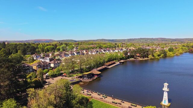 Aerial drone footage of Roath Park in Cardiff, Wales, capturing blue waters of the lake, lakeside greenery and the surrounding residential suburbs under a clear, blue sky. Revealing descending shot.