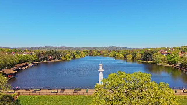 Aerial drone footage of Roath Park in Cardiff, Wales, capturing blue waters of the lake, lakeside greenery and the surrounding residential suburbs under a clear, blue sky. Tracking shot left to right.