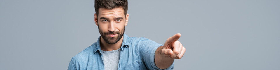 Obraz premium A man with a beard wearing a denim shirt points his finger forward with a serious expression against a neutral gray backdrop.