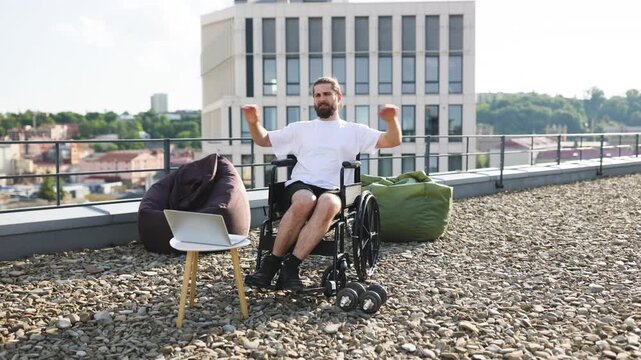 A man in a wheelchair performs exercises on a rooftop terrace with dumbbells and a laptop nearby, overlooking a cityscape