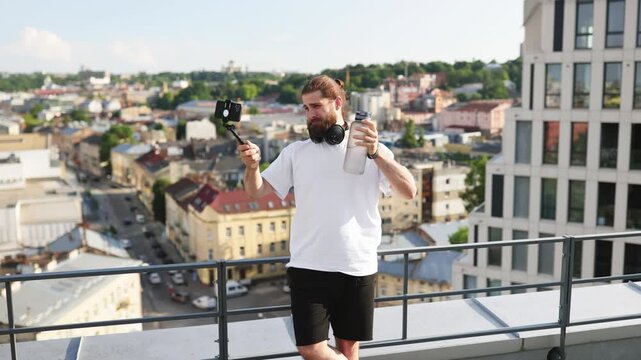 A bearded man with headphones around his neck holds a water bottle and a selfie stick with a phone, recording a video on a rooftop overlooking a cityscape