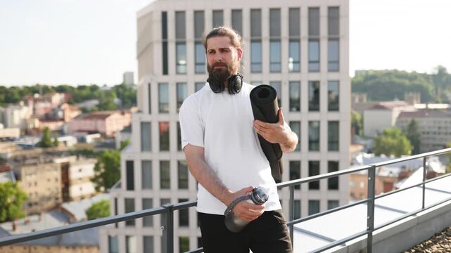 A smiling man with a beard and bun hairstyle holds a rolled-up yoga mat and a water bottle while wearing headphones around his neck on a rooftop with a city view