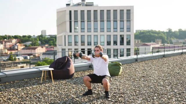A bearded man with long hair performs dumbbell squats on a rooftop terrace with a city view and modern building in the background