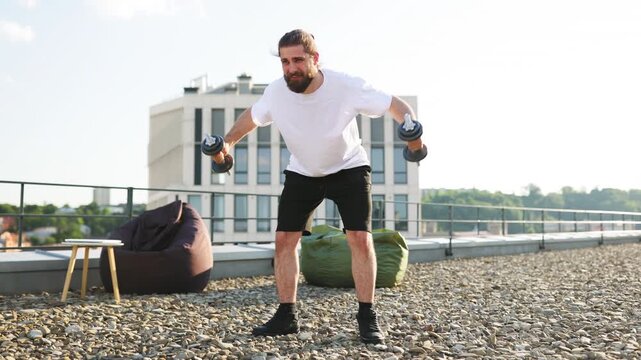 A man with a beard and tied-back hair performs dumbbell lateral raises on a gravel rooftop with a modern building in the background