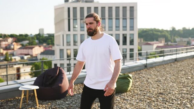 A bearded man in athletic wear stretches his arms and torso while standing on a gravel rooftop with a city view and modern building in the background