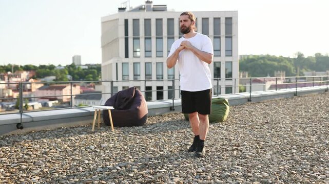 A man with a beard and bun performs a lunge exercise on a gravel rooftop with a modern building in the background