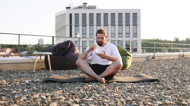 A man with a beard sits cross-legged on a yoga mat on a gravel rooftop, eating a salad from a bowl while a modern building looms in the background