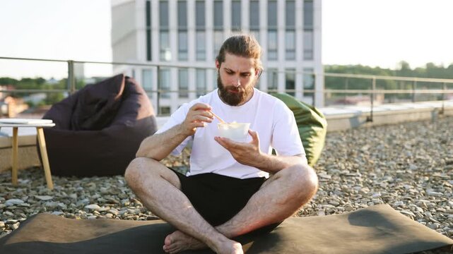 A bearded man with his hair in a bun sits cross-legged on a yoga mat on a rooftop, eating a salad from a container