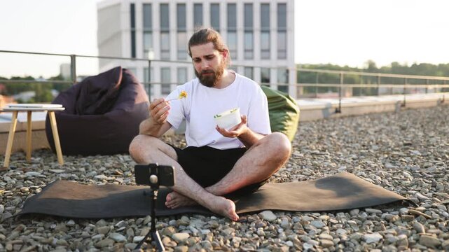 A bearded man sits cross-legged on a yoga mat on a gravel rooftop, eating a salad from a bowl while filming himself on a smartphone