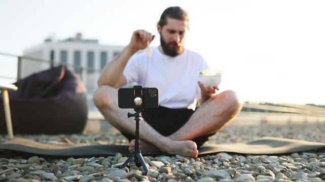 A man sits cross-legged on a yoga mat on a gravel rooftop, eating from a bowl while a smartphone on a tripod records him