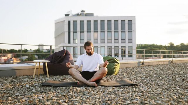 A man with a beard sits in a lotus position on a yoga mat on a gravel rooftop, meditating with his eyes closed while beanbag chairs and a modern building are visible in the background
