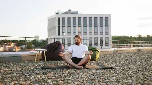 A man with a beard and long hair meditates in a lotus position on a yoga mat on a gravel rooftop with beanbag chairs and a modern building in the background