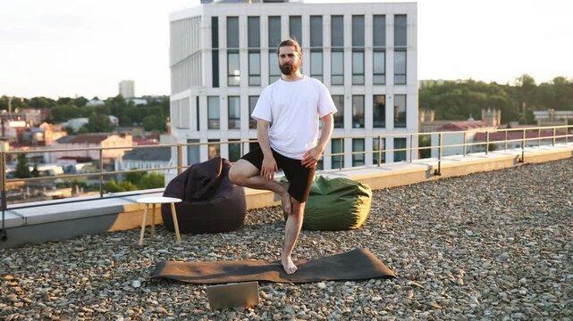 A man with a beard practices yoga in the Vrksasana pose on a rooftop terrace with a city view and beanbag chairs
