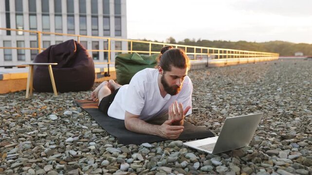 A man with a beard and bun performs a plank exercise on a yoga mat on a rooftop while looking at a laptop