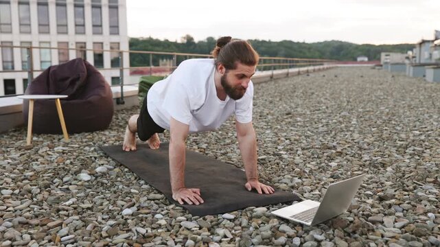 A man with a beard and bun hairstyle performs a pushup on a yoga mat on a gravel rooftop while looking at a laptop