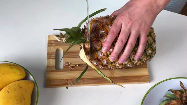 A woman's hands peel a fresh pineapple and cut off the tough stem with a knife on a wooden cutting board. Preparing the tropical fruit for slicing.
