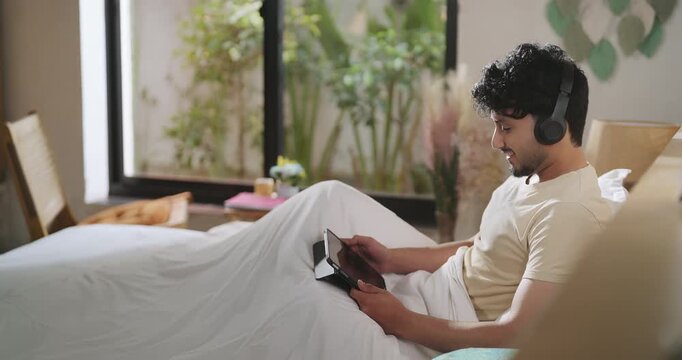 Indian young adult gen z relax man male wear headset sitting on bed use tab look screen listen calm song boy hold tablet play game watch movie film enjoy good mood spend time have fun joy living room