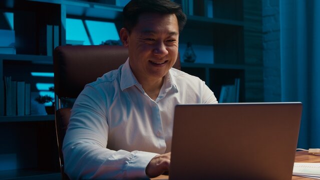 Happy motivated middle-aged Korean business man working laptop in night office Chinese businessman typing computer managing online job task startup Asian male agent Japanese worker work in evening