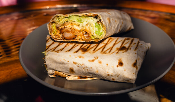 Fresh chicken wrap cut in half on a dark plate. Delicious tortilla filled with grilled meat and vegetable. Tasty street food meal for quick lunch or snack concept in a cafe.