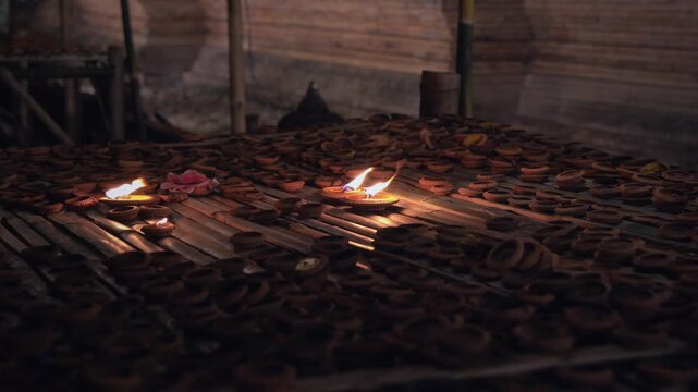 Static wide shot of lit clay oil lamps glowing among hundreds of unlit terracotta diyas on a wooden surface inside a dim rustic structure, warm and devotional atmosphere