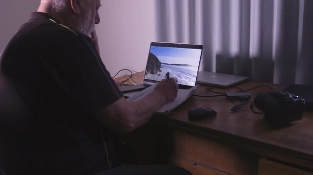 A man sits at a desk, taking notes as he reviews coastal images on his laptop. Suitable for content about remote work or photography editing.