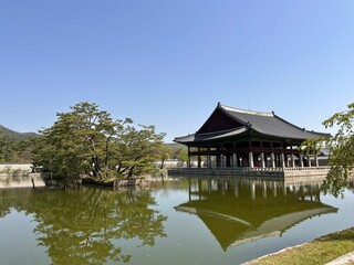 Naklejka premium Bright day by a calm lake with a traditional building reflecting in the water and trees surrounding the area in a historical park
