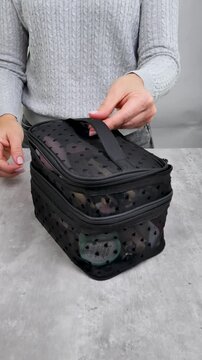A woman putting makeup brushes in a black mesh travel cosmetic bag on a gray table