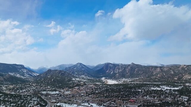 Estes Park mountain range with forested valley and snow patches under cloudy sky, aerial drone overview