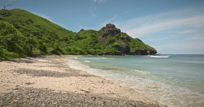 Fiji's Monuriki island beach featuring lush green hills meeting the coastline, with gentle ocean waves lapping the shore under clear blue sky creating a serene tropical island landscape. Drone flight
