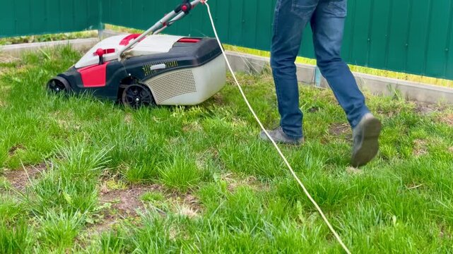 A man in jeans is pushing an electric lawn mower through green grass along a suburban garden fence.