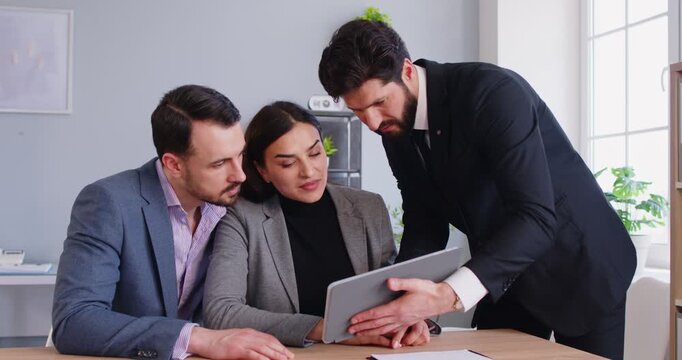Group of people gathered around table, engaged in business discussion, pointing at tablet display screen, focused interaction with computer device, office meeting or consultation, collaborative talk