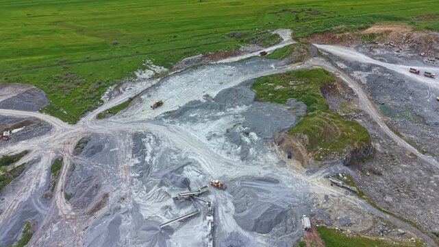 Large equipment moves around in a gravel pit to gather materials. Trucks and machines are present on the rough terrain with green fields in the background under clear skies.