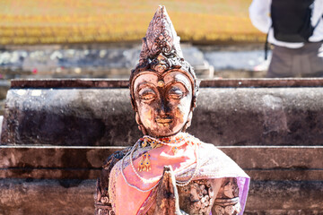 Naklejka premium Weathered female guardian figure stands within a sacred temple site surrounded by historic stone structures and cultural heritage architecture.