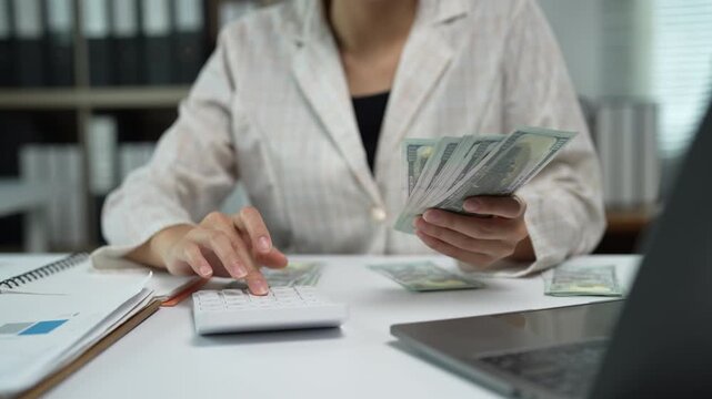 Female accountant or entrepreneur sitting at her office desk, meticulously counting a stack of cash and using a calculator to manage finances, budget expenses, and calculate profits