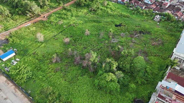 A view from above of an urban vacant lot overgrown with weeds and surrounded by buildings and greenery in Indonesia.