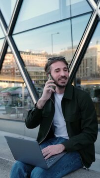 Businessman sitting outdoors with a laptop and smartphone, engaging in a business conversation