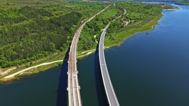 Two bridges run parallel over a river with clear water. Lush green hills and trees surround the area. The view shows a blend of nature and infrastructure under a bright sky.