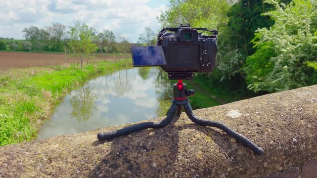Mirrorless camera recording nature timelapse on flexible travel tripod. Outdoor videography equipment overlooking river landscape under sunny countryside sky. Content creation filmmaking technology in