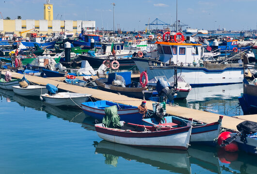 Colorful traditional fishing boats moored in the calm, reflective water of Setubal harbor, Portugal. Stone steps and a beached boat on the shore. Setubal, Portugal. 