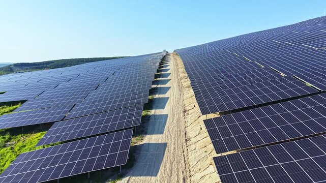 Rows of solar panels stretch across a hillside, collecting sunlight. The landscape shows a clear blue sky with no clouds. The panels are aligned for maximum sun exposure.