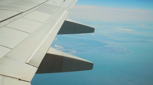 Airplane flying above coastal area with visible wing and flap support fairings. Jet cruising over shoreline showing aerodynamic structures and control panels. Aircraft soaring through sky displaying