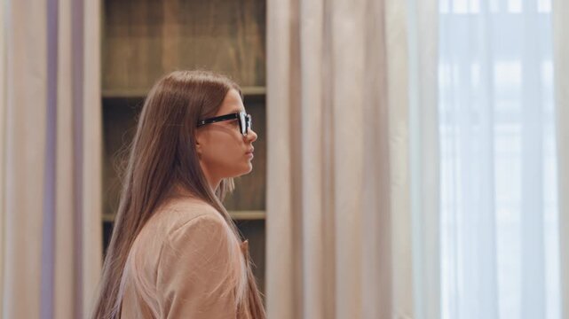 Woman with glasses inspects hotel lobby. Longhaired woman in trench coat slowly surveys chandelier, curtains, and bookcase in warmtoned boutique lobby soft daylight through drapes, velvet seating,