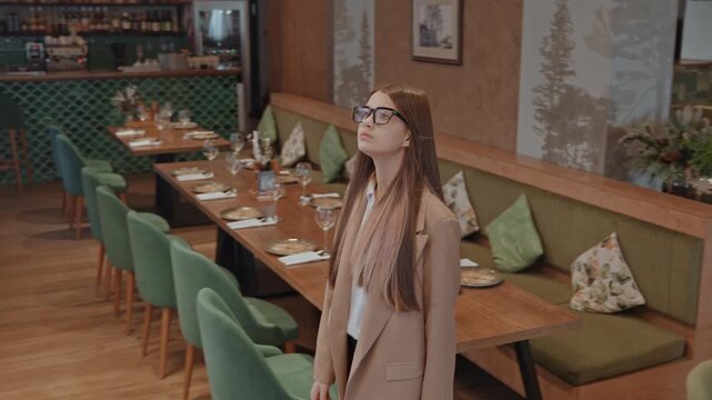 Confident white woman exploring restaurant space, glasses and beige coat, purposeful stride between tables, checking seating layout and table settings, placing laptop bag on chair, reviewing documents