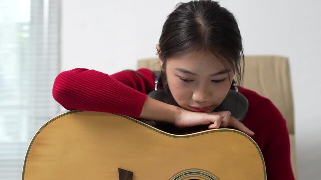 Thoughtful young asian musician feeling sad and melancholic while resting on her acoustic guitar, struggling with writer's block and lack of creative inspiration