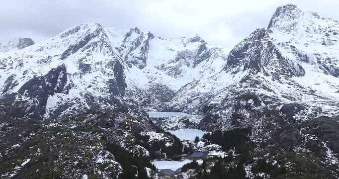 Flakstad&oslash;ya mountain ridge near Nusfjord in Lofoten Islands shows frozen lakes in glacial basins between granite peaks, revealing alpine terrain as drone slides right and pans and reveal